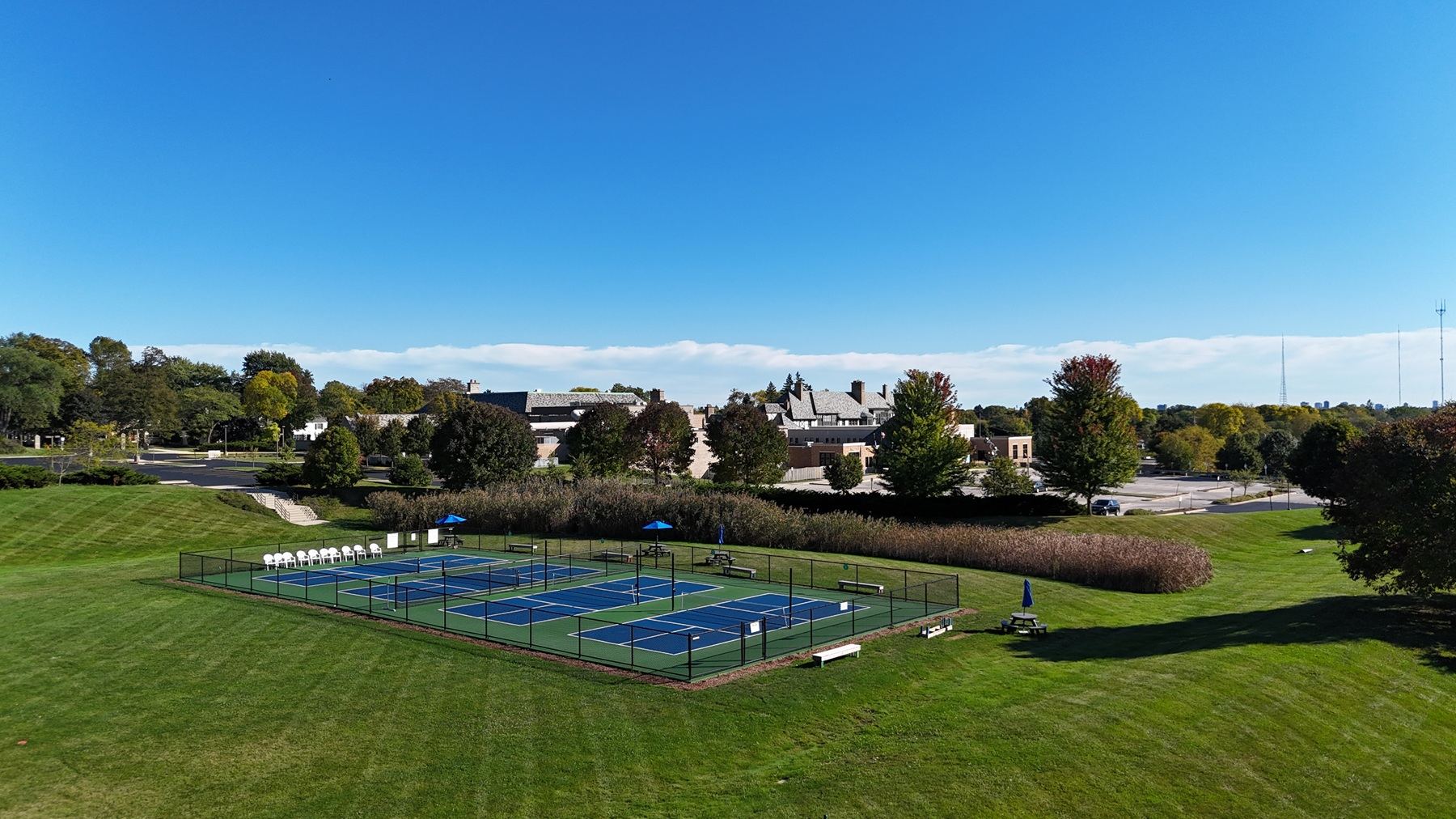Four fenced pickleball courts on a rolling lawn with a building and trees beyond