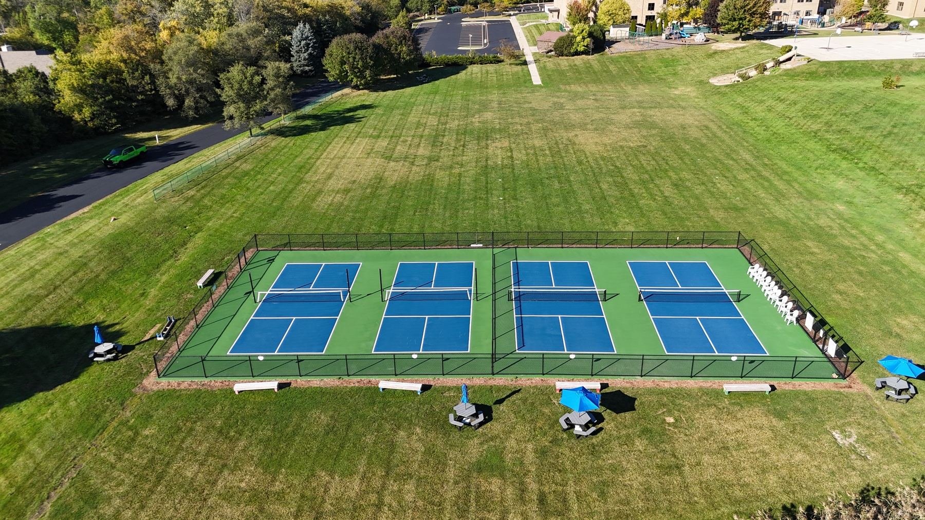 Overhead view of four blue pickleball courts on a green surround, enclosed by black chain-link fence with a center divider, benches and umbrella tables outside