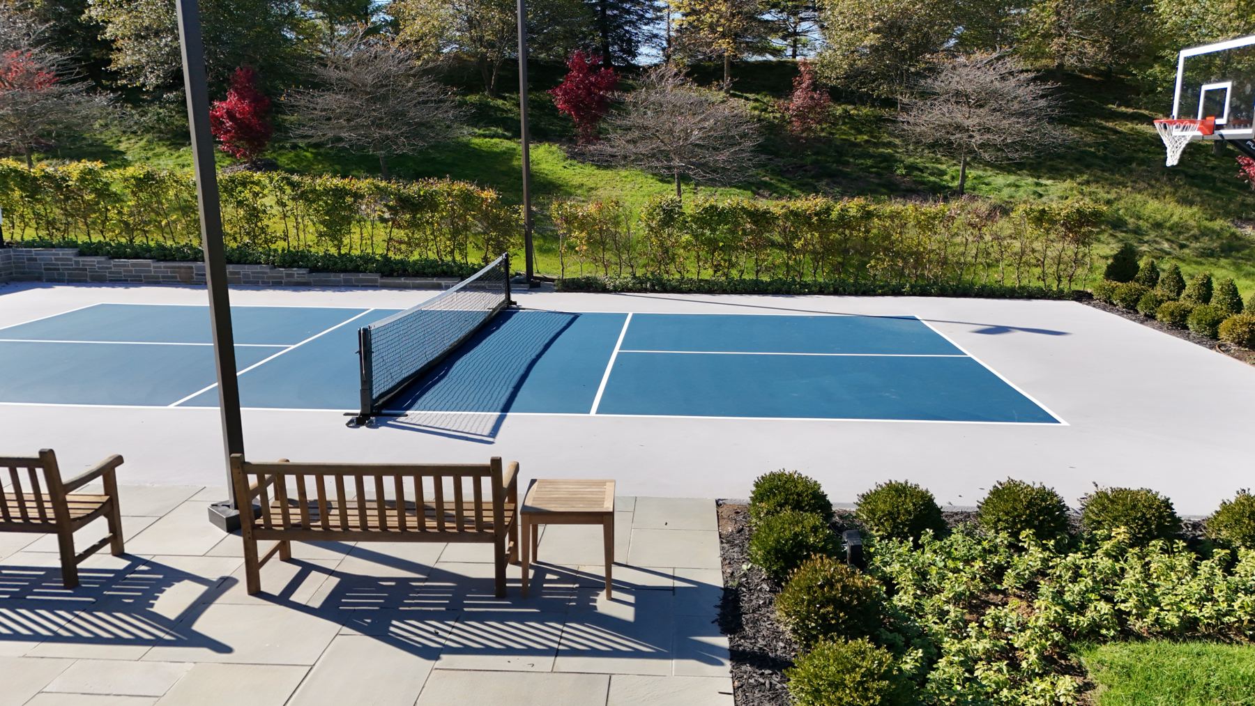 View from a bluestone patio with teak benches across a blue pickleball court to a hedge and stone wall