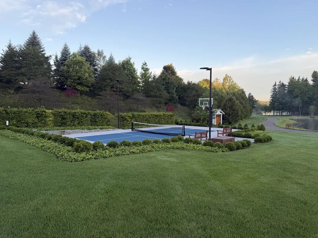 Private blue pickleball court framed by clipped hedges, lawn and mature trees beside a pond